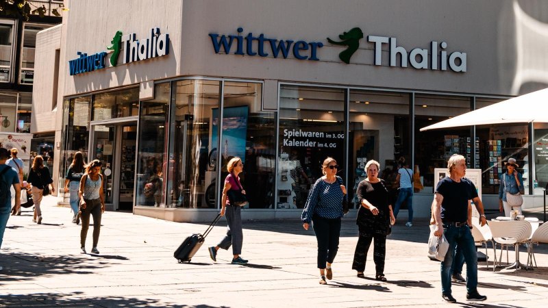 Menschen flanieren vor der Buchhandlung Wittwer-Thalia. Einige sitzen unter Sonnenschirmen in einem Café. Es ist ein sonniger Tag., © Stuttgart Marketing GmbH, Sarah Schmid Menschen flanieren vor der Buchhandlung Wittwer-Thalia. Einige sitzen unter Sonnenschirmen in einem Café. Es ist ein sonniger Tag., © Stuttgart Marketing GmbH, Sarah Schmid