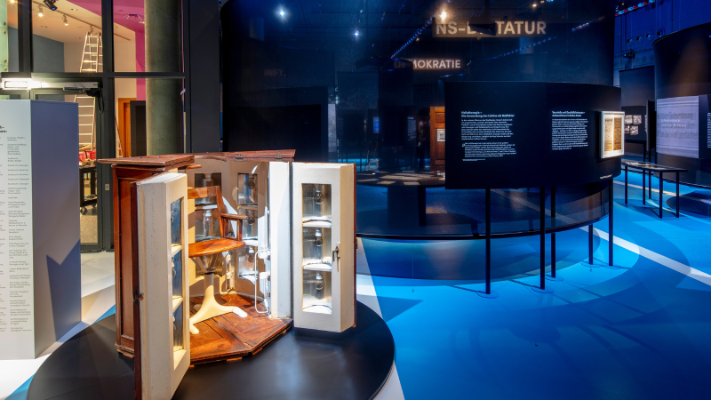 Ein moderner Ausstellungsraum mit blauen Böden, Texttafeln und einem geöffneten Holzschrank mit Stuhl. Im Hintergrund sind weitere Exponate zu sehen., © Frei_Schwimmen_Daniel_Stauch