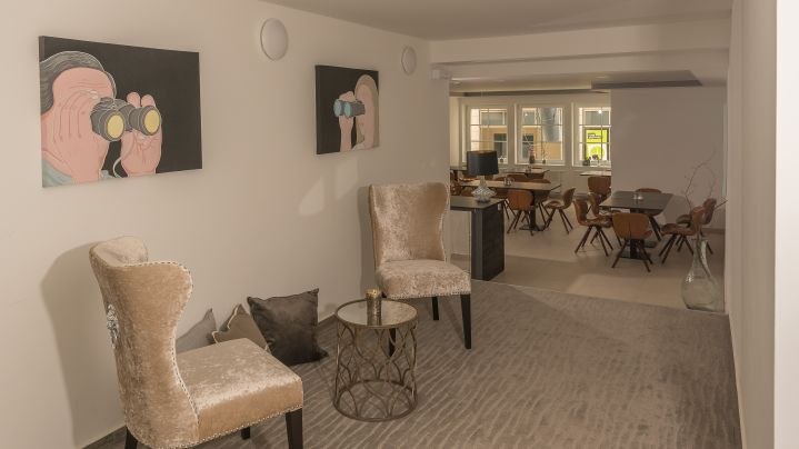 Elegante Lobby mit zwei beigen Sesseln, kleinem Tisch und Kunstwerken an der Wand. Im Hintergrund ein Raum mit Tischen und Stühlen., © Kronenstuben Boutique-Hotel