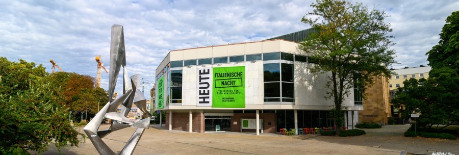 Modern sculpture in front of the Schauspielhaus with large windows and posters for 'Italian Night'. Cloudy sky, trees and construction cranes in the background., © Björn Klein Modern sculpture in front of the Schauspielhaus with large windows and posters for 'Italian Night'. Cloudy sky, trees and construction cranes in the background., © Björn Klein