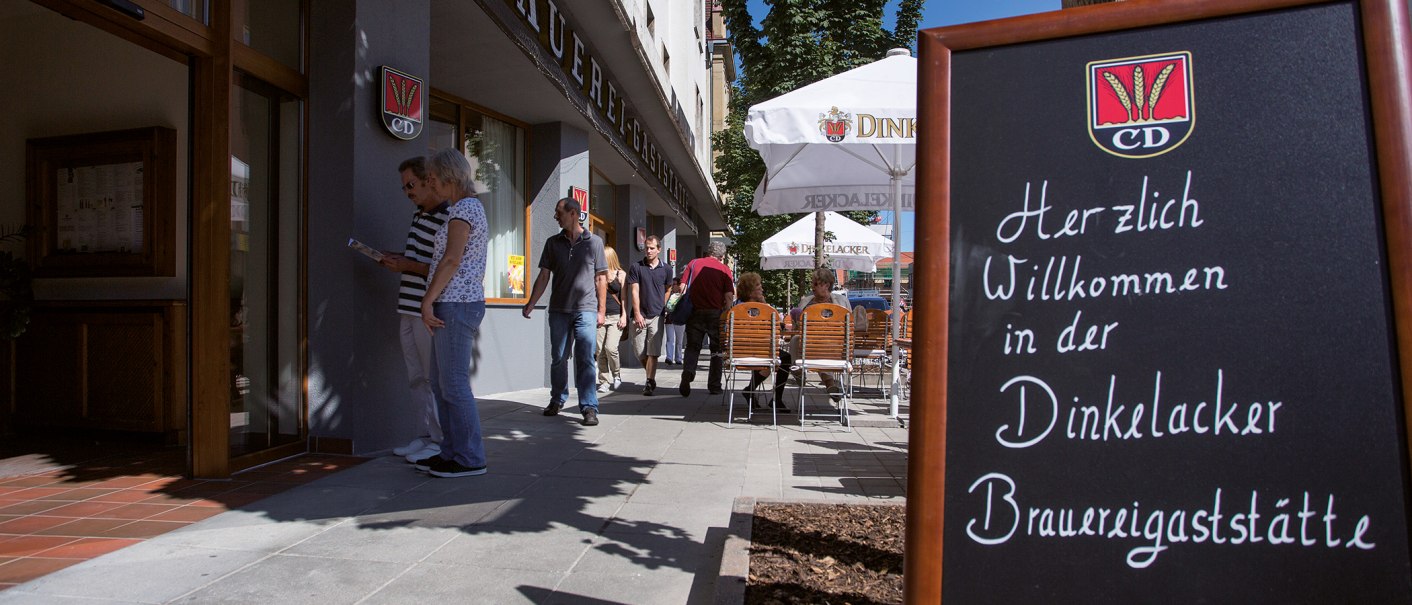 Eingang der Dinkelacker Brauereigaststätte mit einem Willkommensschild. Menschen gehen vorbei, einige sitzen an Tischen unter Sonnenschirmen., © Familienbrauerei Dinkelacker Eingang der Dinkelacker Brauereigaststätte mit einem Willkommensschild. Menschen gehen vorbei, einige sitzen an Tischen unter Sonnenschirmen., © Familienbrauerei Dinkelacker