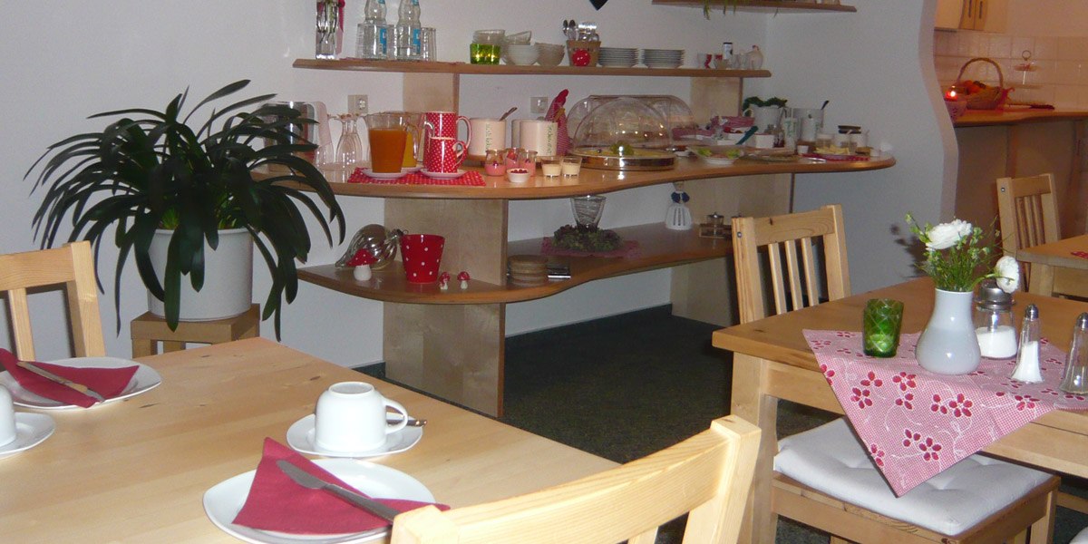 Ein gemütlicher Frühstücksraum mit Holztischen, roten Servietten und einem Buffet mit Getränken und Speisen. Eine Pflanze steht im Raum., © Hotel Hochwald