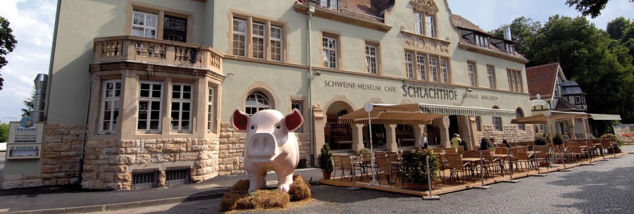 Historic building with pig museum and abattoir café. Large pig figure stands in front of it, surrounded by tables and chairs in the outdoor area., © Buschwerk Mediendesign-Wilhelmer Gastronomie