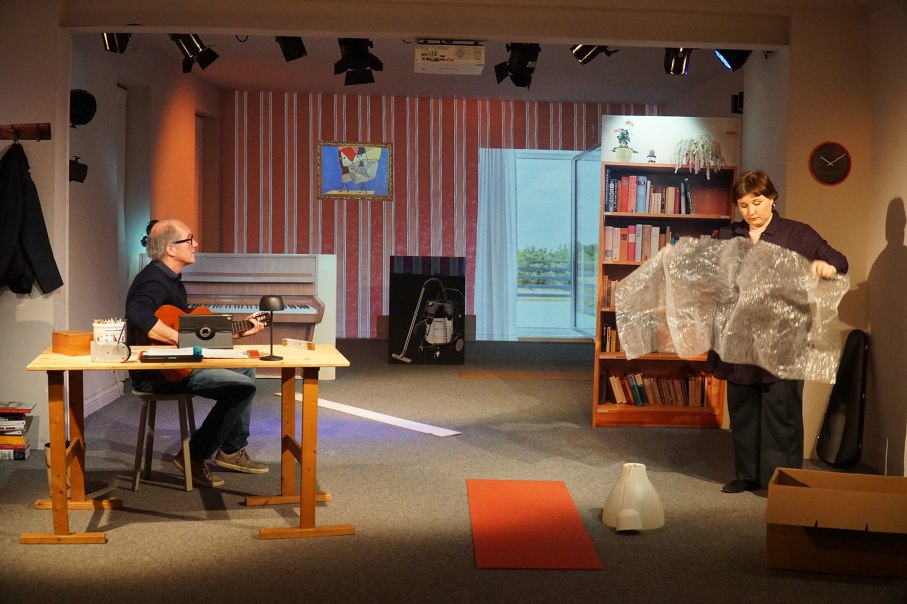 Ein Mann spielt Gitarre an einem Tisch, während eine Frau Luftpolsterfolie hält. Im Hintergrund sind ein Bücherregal und ein Klavier zu sehen., © Katja Ritter Ein Mann spielt Gitarre an einem Tisch, während eine Frau Luftpolsterfolie hält. Im Hintergrund sind ein Bücherregal und ein Klavier zu sehen., © Katja Ritter