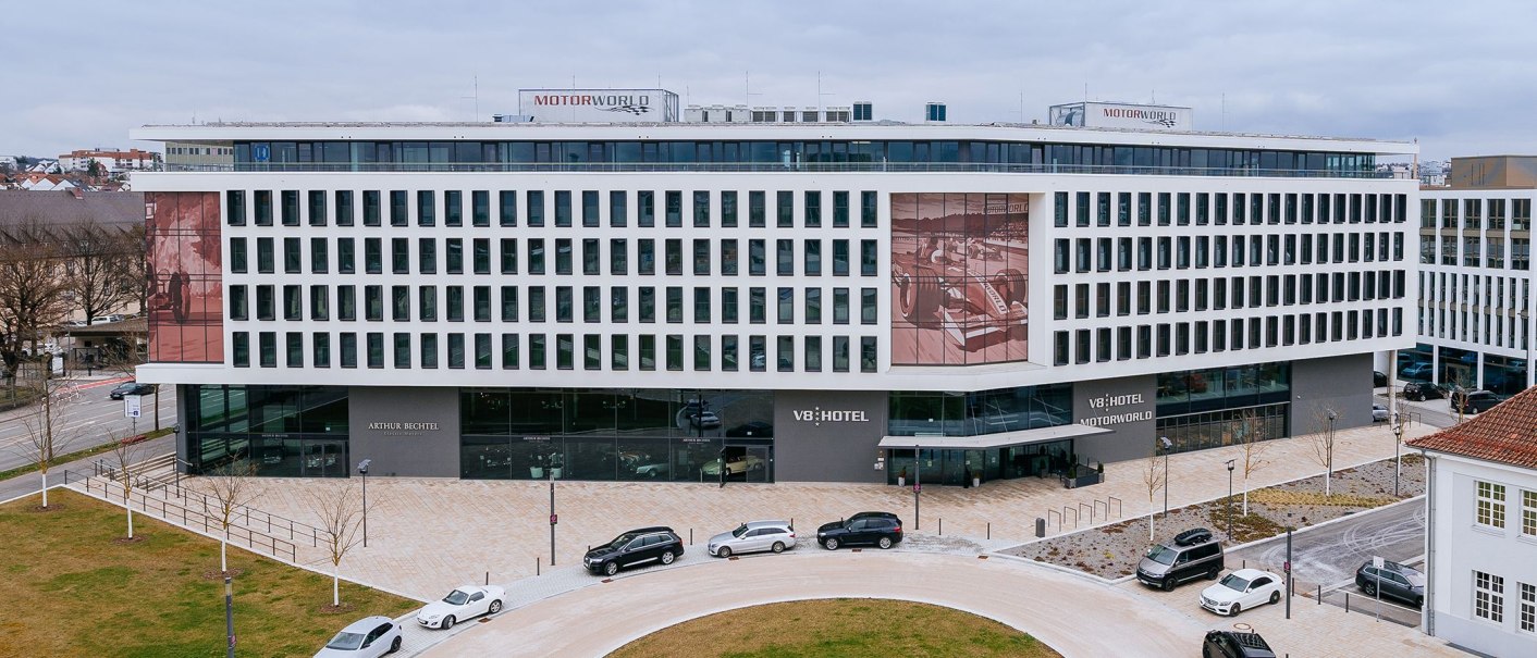Das V8 HOTEL ist ein modernes Geb&auml;ude mit vielen Fenstern. Vor dem Hotel parken mehrere Autos. Auf dem Dach steht "MOTORWORLD"., &copy; V8 HOTEL