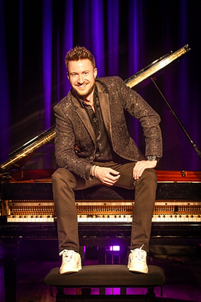 A man in a glittering jacket sits smiling on a piano in front of purple curtains., &copy; Renitenztheater Stuttgart e.V.