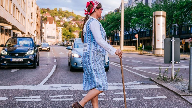 Eine Frau mit Kopftuch und Besen überquert eine Straße in einer Stadt. Autos sind im Hintergrund zu sehen., © Thomas Niedermüller Eine Frau mit Kopftuch und Besen überquert eine Straße in einer Stadt. Autos sind im Hintergrund zu sehen., © Thomas Niedermüller