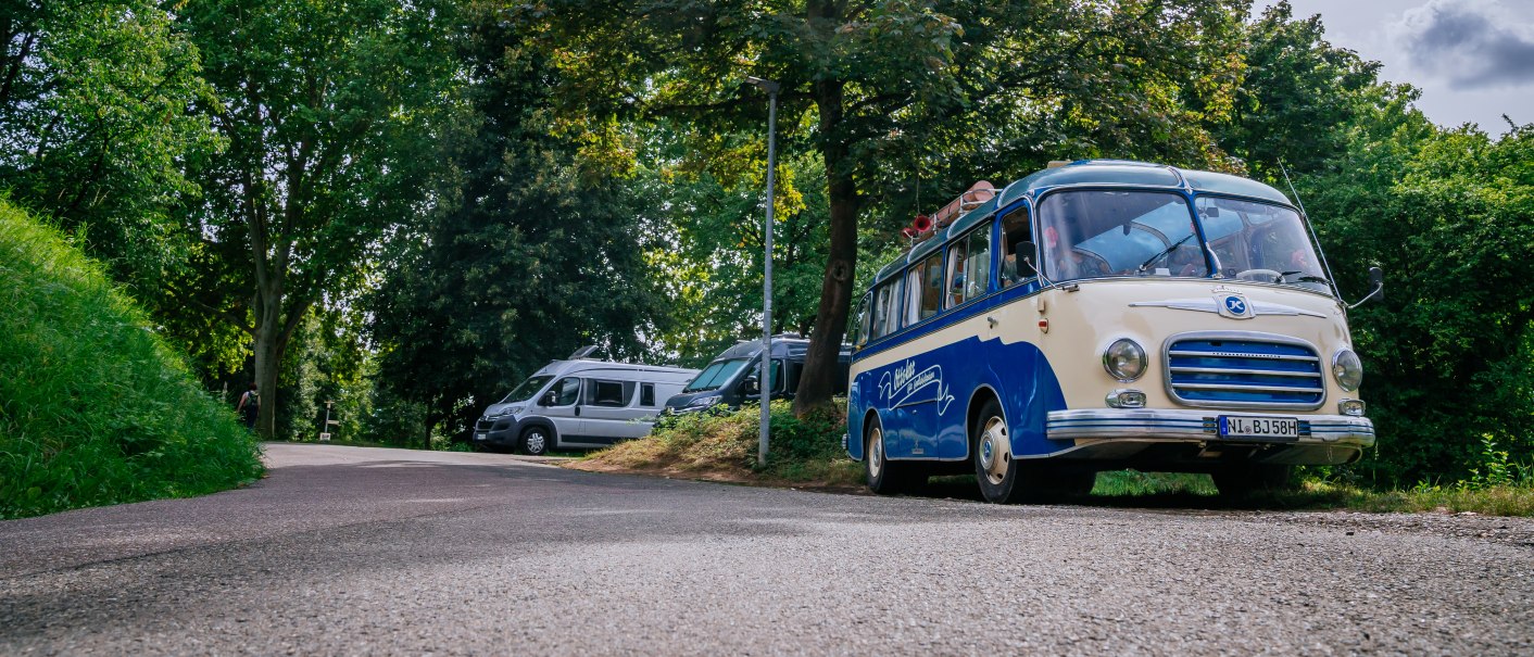 Ein blauer Oldtimer-Bus steht auf einem Wohnmobilstellplatz neben einem modernen Wohnmobil, umgeben von grünen Bäumen und blauem Himmel., © Stuttgart-Marketing GmbH, Thomas Niedermüller Ein blauer Oldtimer-Bus steht auf einem Wohnmobilstellplatz neben einem modernen Wohnmobil, umgeben von grünen Bäumen und blauem Himmel., © Stuttgart-Marketing GmbH, Thomas Niedermüller