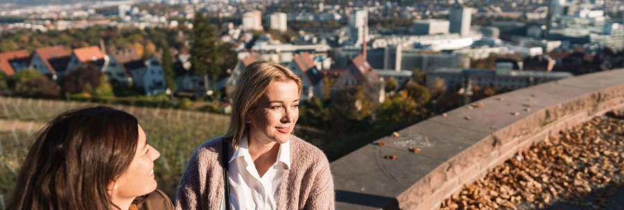 Zwei Frauen sitzen auf einer Mauer, genießen die Aussicht auf eine Stadt mit Hügeln im Hintergrund. Herbstlaub liegt auf der Mauer., © SMG, wpsteinheisser Zwei Frauen sitzen auf einer Mauer, genießen die Aussicht auf eine Stadt mit Hügeln im Hintergrund. Herbstlaub liegt auf der Mauer., © SMG, wpsteinheisser