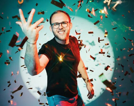 Man with glasses and beard in front of confetti and the lettering 'SVENOMENAL'. He is wearing a black T-shirt and smiling at the camera., &copy; Theaterhaus Stuttgart e.V.