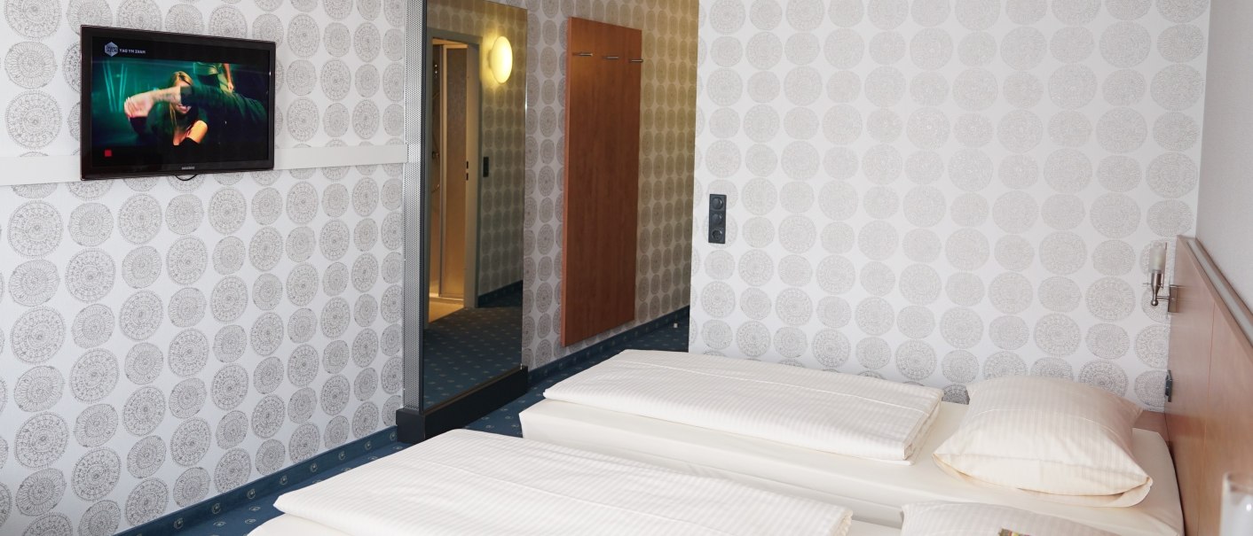 A hotel room with two single beds, patterned wallpaper, a wall-mounted TV, and a mirror. A hallway leads to a well-lit area., &copy; TOMAS
