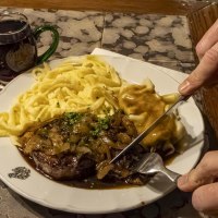 Ein Teller mit Zwiebelrostbraten, Sp&auml;tzle und So&szlig;e. Daneben stehen zwei Gl&auml;ser mit Getr&auml;nken. Eine Person schneidet das Fleisch., &copy; Stuttgart-Marketing GmbH, Sarah Schmid