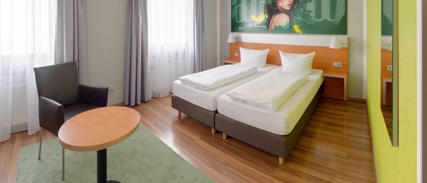 Modern hotel room with two single beds, armchair, table and large mural. Light-colored curtains and wooden floor create an inviting atmosphere., © attimo Hotel Stuttgart Modern hotel room with two single beds, armchair, table and large mural. Light-colored curtains and wooden floor create an inviting atmosphere., © attimo Hotel Stuttgart
