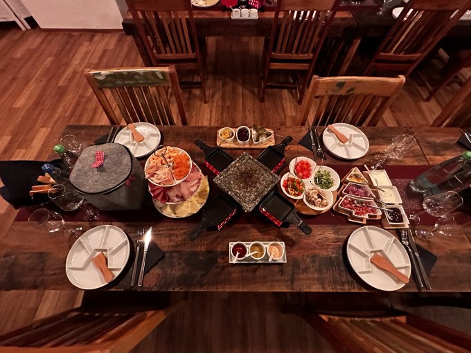 Ein Tisch mit Raclette-Grill, Tellern, Besteck und verschiedenen Zutaten für einen Raclette-Abend, darunter Gemüse, Fleisch und Saucen., © Cool-Tours StattReisen Ein Tisch mit Raclette-Grill, Tellern, Besteck und verschiedenen Zutaten für einen Raclette-Abend, darunter Gemüse, Fleisch und Saucen., © Cool-Tours StattReisen