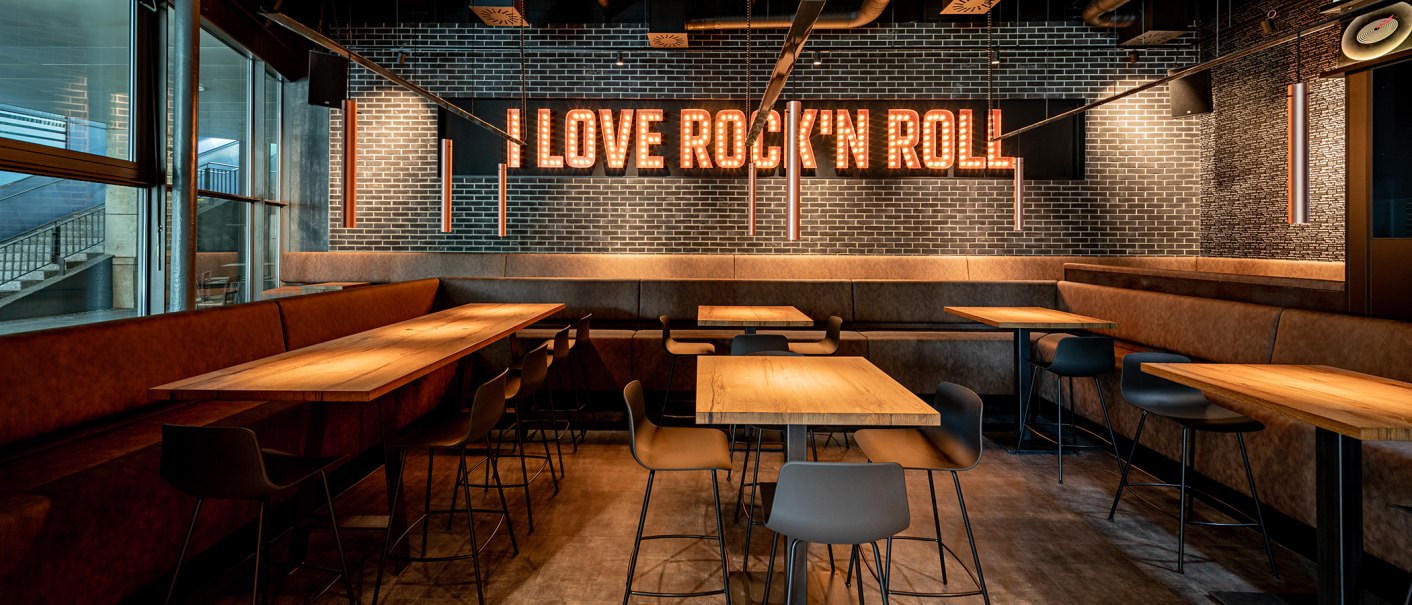 Ein stilvolles Café mit Holztischen und modernen Stühlen. An der Wand leuchtet der Schriftzug 'I Love Rock 'n Roll' in großen Buchstaben., © Maks Richter