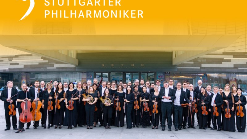 Die Stuttgarter Philharmoniker posieren mit ihren Instrumenten vor einem modernen Geb&auml;ude. Ein gelbes Banner mit ihrem Namen ist oben zu sehen., &copy; Juan Carlos Rivas Perretta, Stuttgarter Philharmoniker