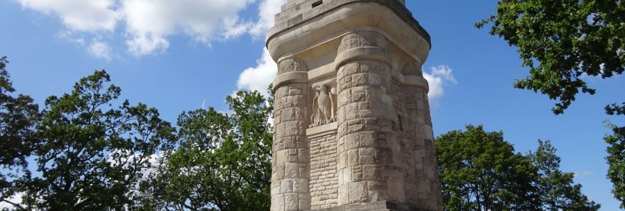 Ein massiver Bismarckturm aus Stein steht auf einem Platz, umgeben von Bäumen und unter einem blauen Himmel mit wenigen Wolken., © SMG Ein massiver Bismarckturm aus Stein steht auf einem Platz, umgeben von Bäumen und unter einem blauen Himmel mit wenigen Wolken., © SMG