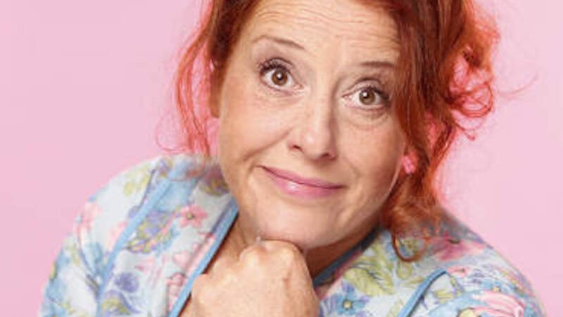 A woman with red hair and a floral top against a pink background, smiling and resting her hand on her chin., © Renitenztheater Stuttgart e.V. A woman with red hair and a floral top against a pink background, smiling and resting her hand on her chin., © Renitenztheater Stuttgart e.V.