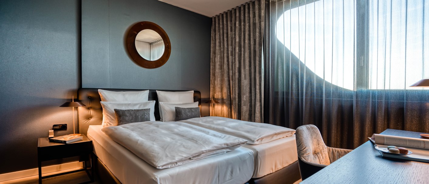 Modernes Hotelzimmer mit Doppelbett, rundem Spiegel an der Wand und großem Fenster mit Vorhängen. Ein Schreibtisch mit Stuhl steht daneben., © CLOUD N°7 APARTMENTS GmbH