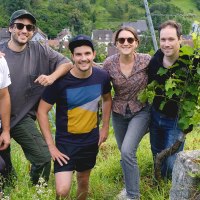 Fünf Personen posieren lächelnd in einem Weinberg. Im Hintergrund ist ein Dorf zu sehen. Die Gruppe wirkt entspannt und naturverbunden., © Weinkollektiv Edenberg & Tal Fünf Personen posieren lächelnd in einem Weinberg. Im Hintergrund ist ein Dorf zu sehen. Die Gruppe wirkt entspannt und naturverbunden., © Weinkollektiv Edenberg & Tal