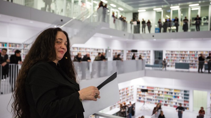 Eine Frau mit langen Haaren und Mikrofon steht in einer modernen, mehrstöckigen Bibliothek mit vielen Zuschauern auf den Galerien., © Foto: die arge lola