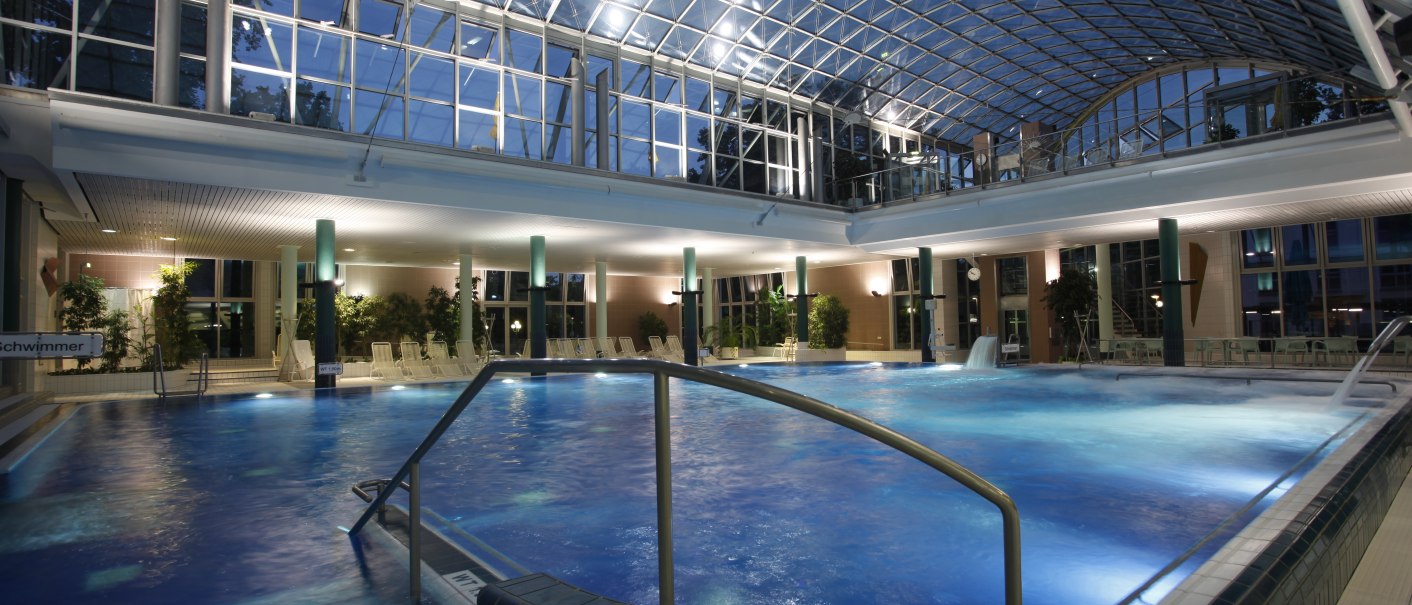 Beleuchtetes Schwimmbad mit Glasdach und Pflanzen. Der Pool ist von Liegestühlen umgeben, die Atmosphäre ist ruhig und entspannend., © Stuttgarter Bäder