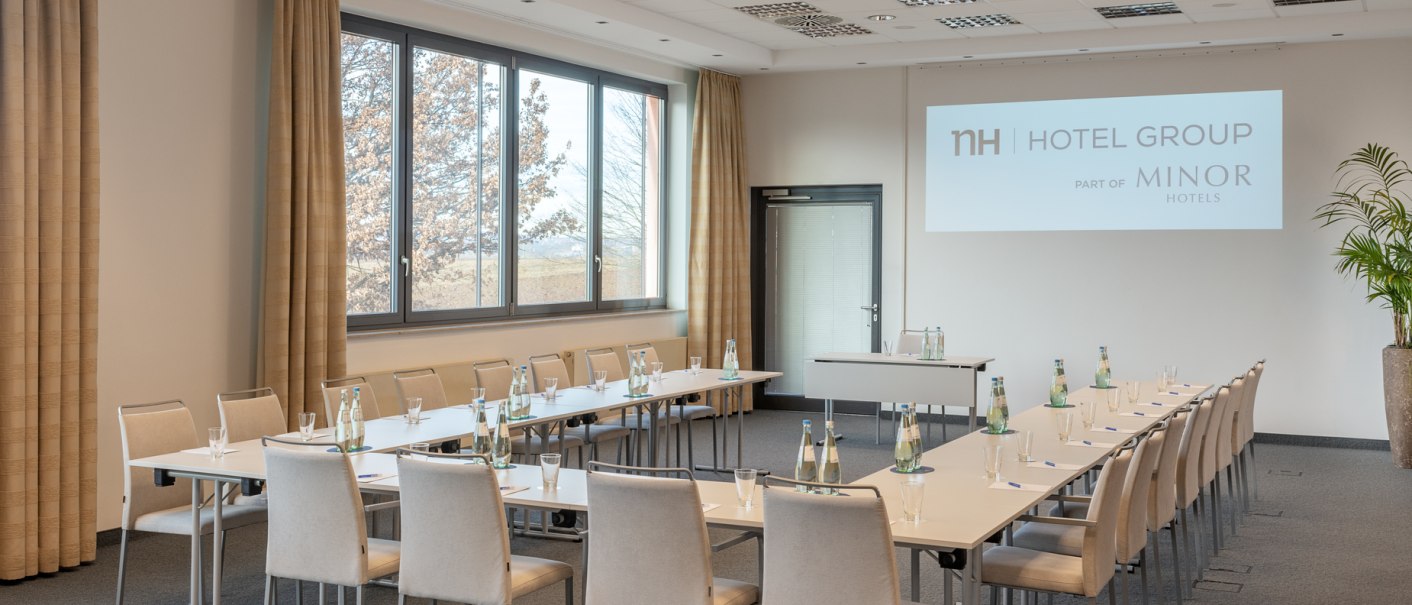 Moderner Konferenzraum mit U-förmiger Tischanordnung, Stühlen, Wasserflaschen und Gläsern. An der Wand wird das Logo der NH Hotel Group projiziert., © NH Hotel Stuttgart Airport Moderner Konferenzraum mit U-förmiger Tischanordnung, Stühlen, Wasserflaschen und Gläsern. An der Wand wird das Logo der NH Hotel Group projiziert., © NH Hotel Stuttgart Airport