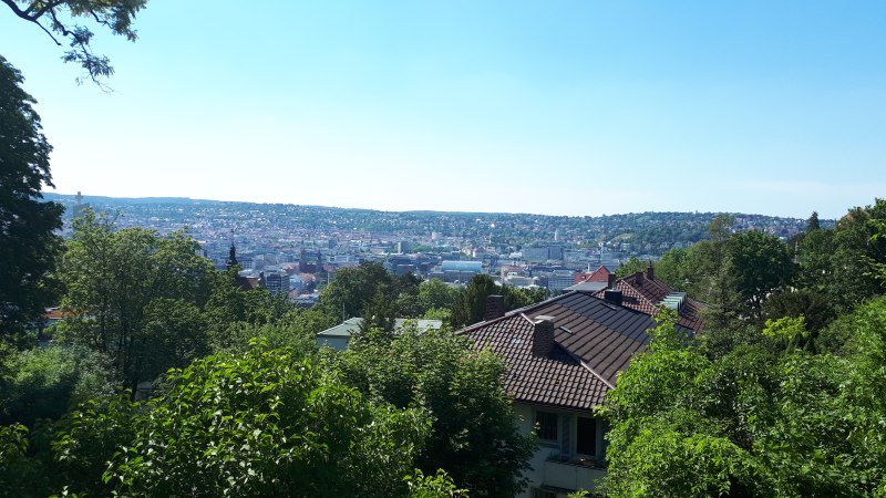 Panoramablick auf eine Stadt mit gr&uuml;nen B&auml;umen im Vordergrund und H&uuml;geln im Hintergrund unter klarem Himmel., &copy; SMG