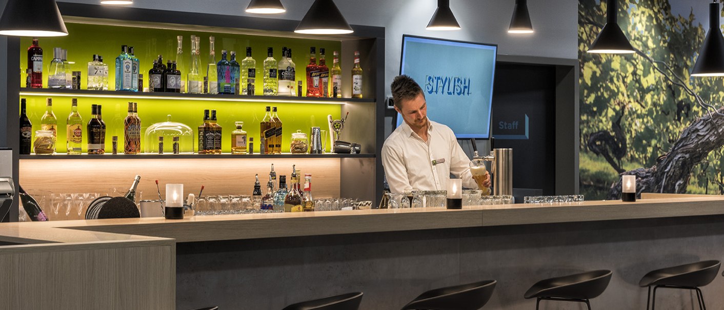 Ein Barkeeper zapft ein Bier an einer stilvollen Bar mit beleuchteten Flaschenregalen und modernen Hockern., © ibis Styles Stuttgart Vaihingen