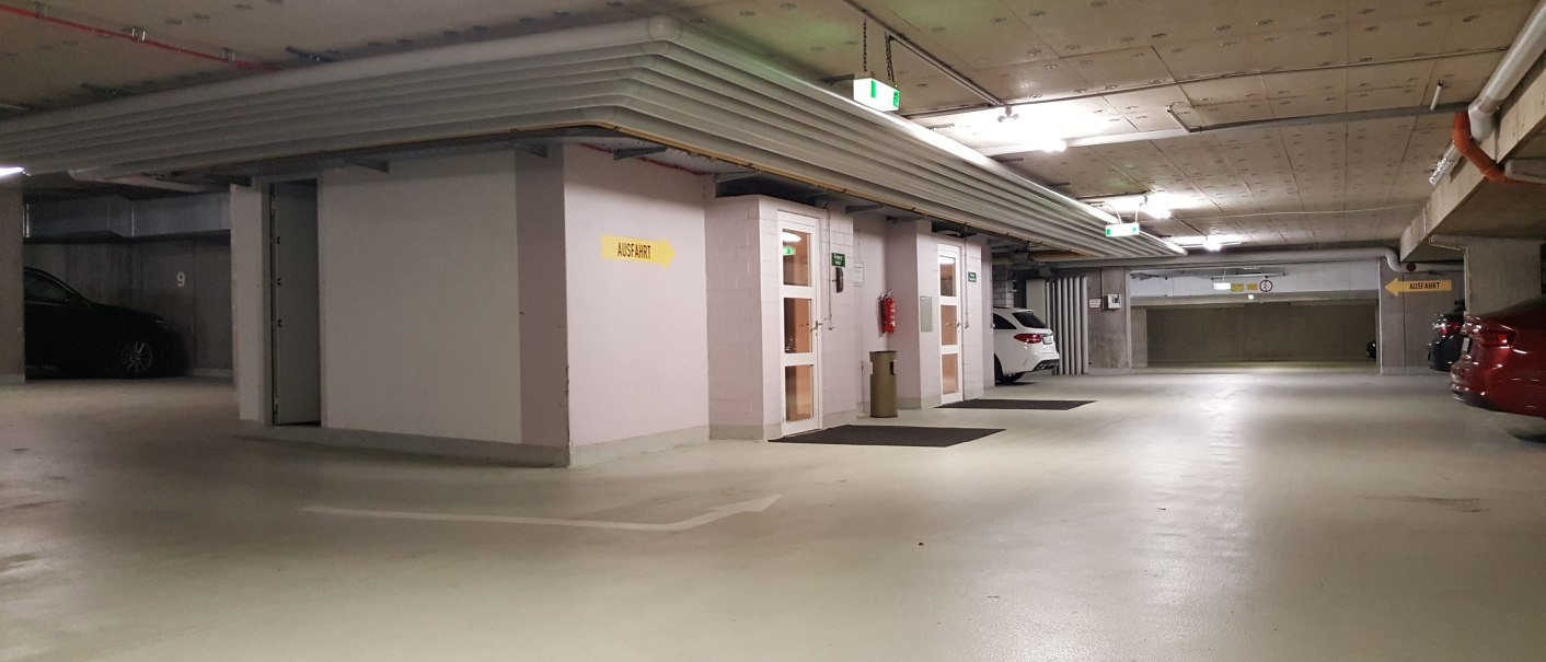 Underground parking lot with cars, exit signs and lighting. White and red vehicles are visible, as well as a yellow exit sign., © Erikson Hotel