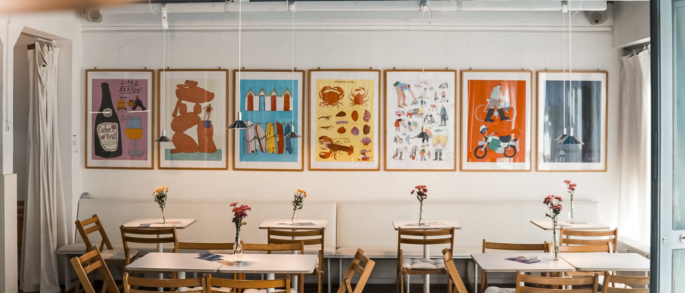 A cosy café with wooden furniture, colorful posters on the wall and flowers on the tables. A projector hangs from the ceiling., © Merlin, Stuttgart