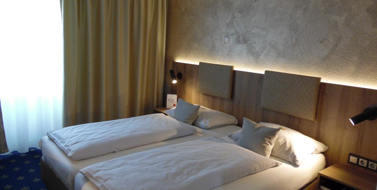 Modernes Doppelzimmer mit zwei Einzelbetten, Nachttischlampen und einem Fenster mit Vorhängen. Die Wand ist mit Holzpaneelen verkleidet., © Hotel Arcis