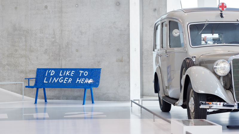 Blue bench with the inscription 'I'd like to linger here' next to a classic car in a modern showroom., © Finnegan Shannon Blue bench with the inscription 'I'd like to linger here' next to a classic car in a modern showroom., © Finnegan Shannon