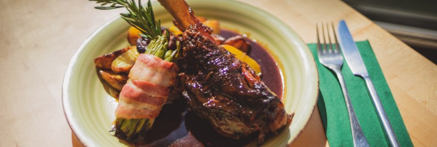 A plate with leg of lamb, asparagus wrapped in bacon and potatoes in sauce. Next to it are cutlery and a green napkin., © Rotenberger Weingärtle, Frederik Garlin