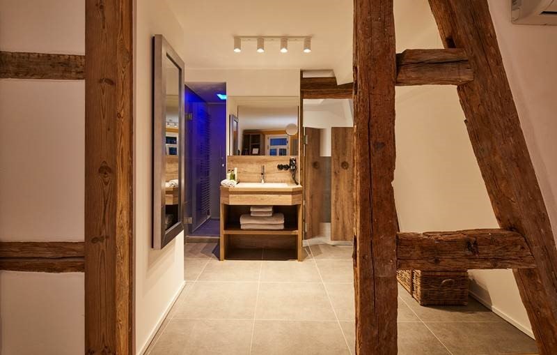 Modernes Badezimmer mit sichtbaren Holzbalken, einem Waschtisch mit Spiegel und Handt&uuml;chern. Beleuchtung und blauer Lichtakzent im Hintergrund., &copy; Hotel Restaurant Bachofer