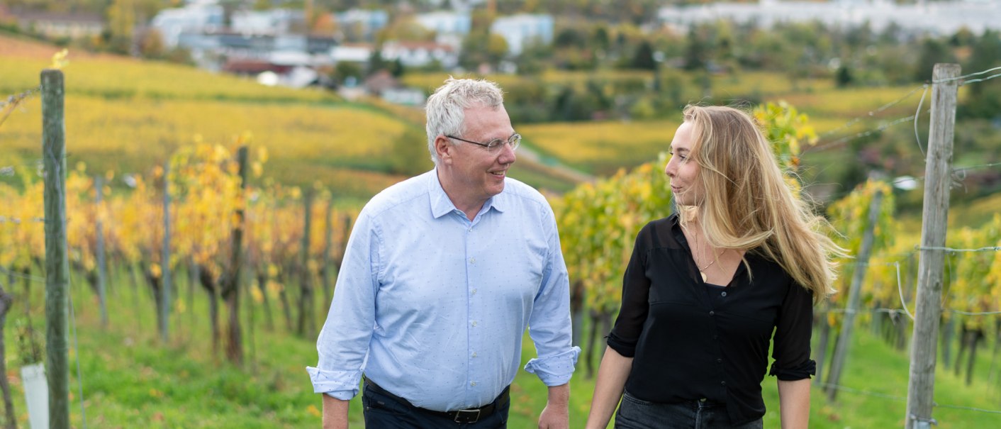 Zwei Personen spazieren l&auml;chelnd durch einen herbstlichen Weinberg. Im Hintergrund sind gelbe Weinreben und eine unscharfe Landschaft zu sehen., &copy; BURKHARDT HELLWIG