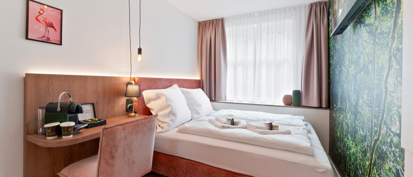 Modernes Zimmer mit Doppelbett, rosa Akzenten, Wandbild eines Waldes und Flamingo-Bild. Schreibtisch mit Kaffeemaschine und stilvoller Beleuchtung., &copy; JUNGLE N&deg;5 GmbH