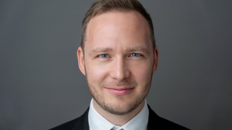 A man in a suit smiles at the camera. The background is gray., &copy; Renitenztheater Stuttgart e.V.