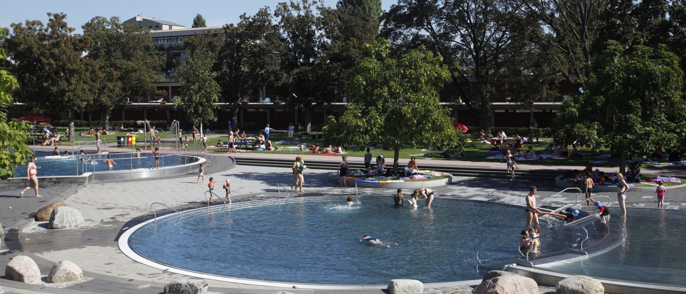 Das Inselbad Stuttgart-Untertürkheim zeigt Menschen, die an einem sonnigen Tag in Pools schwimmen und auf grünen Wiesen entspannen., © Stuttgarter Bäder