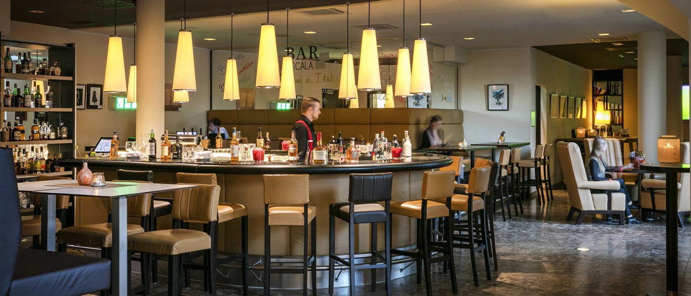 Elegant hotel bar with round counter, bar stools and subdued lighting. Guests sit at tables while a bartender works behind the counter., © Arcotel Hotels Elegant hotel bar with round counter, bar stools and subdued lighting. Guests sit at tables while a bartender works behind the counter., © Arcotel Hotels