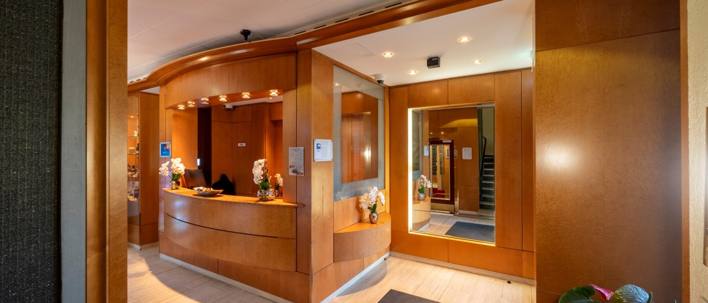 Elegant hotel reception with wood paneling, flowers and mirror. Warm lighting creates an inviting atmosphere., © PLAZA INN Rieker Stuttgart Hauptbahnhof