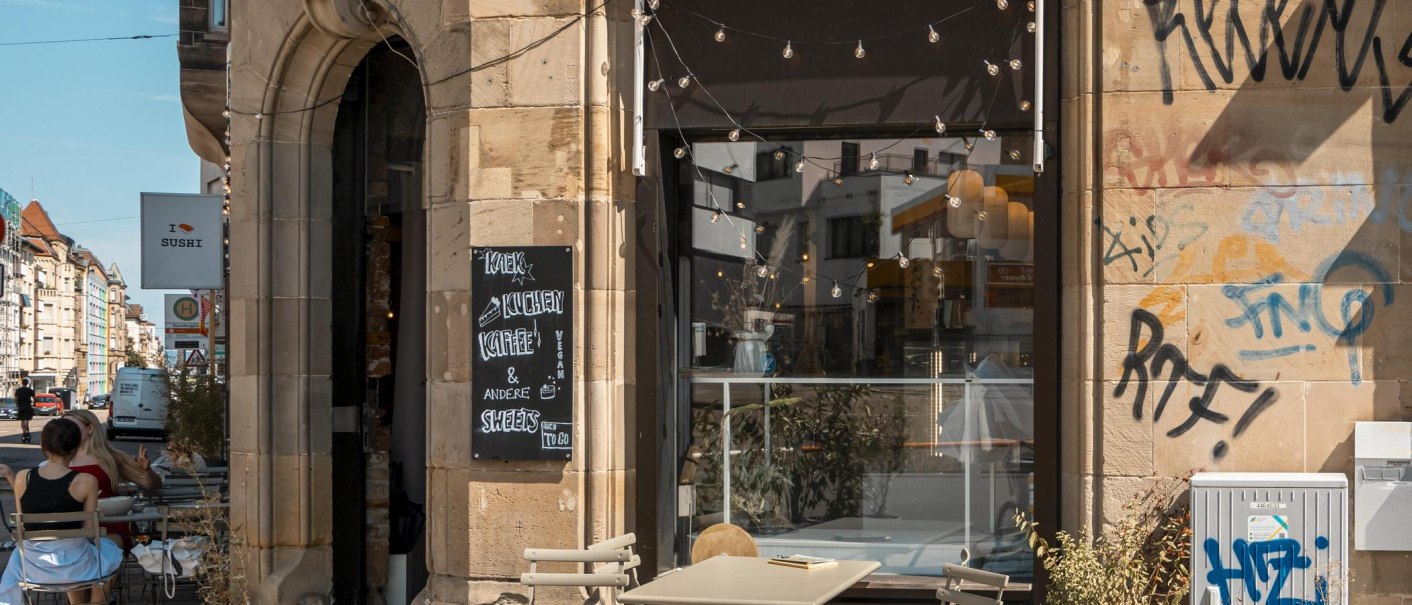 Café mit Außenbestuhlung, Graffiti an der Wand, Lichterketten über dem Eingang. Schild mit "Kaffee & Kuchen". Stadtstraße im Hintergrund., © SMG, Sarah Schmid Café mit Außenbestuhlung, Graffiti an der Wand, Lichterketten über dem Eingang. Schild mit "Kaffee & Kuchen". Stadtstraße im Hintergrund., © SMG, Sarah Schmid
