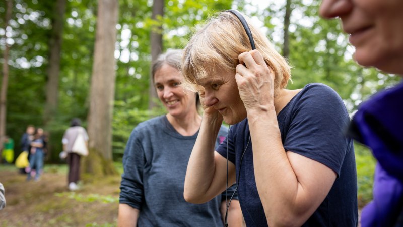 Zwei Frauen im Wald, eine h&ouml;rt mit Kopfh&ouml;rern zu, w&auml;hrend die andere l&auml;chelt. Im Hintergrund sind B&auml;ume und weitere Personen zu erkennen., &copy; Julian Rettig