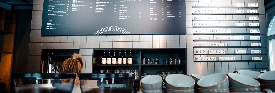 Café interior with coffee bean containers in the foreground and drinks menu on the wall. A person is standing behind the counter., © SMG Stuttgart Marketing GmbH - Sarah Schmid