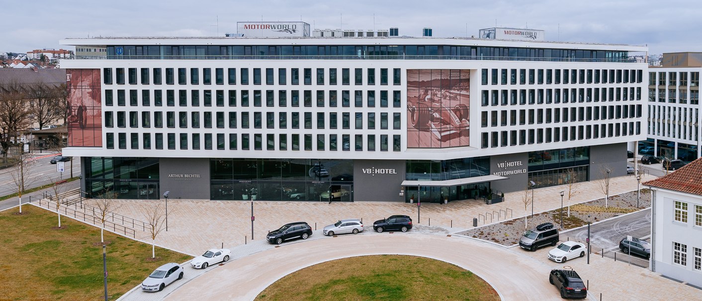 Moderne Fassade des V8 Hotels mit großen Fenstern und parkenden Autos davor. Auf dem Dach ist das Motorworld-Logo sichtbar., © Thomas Niedermüller Moderne Fassade des V8 Hotels mit großen Fenstern und parkenden Autos davor. Auf dem Dach ist das Motorworld-Logo sichtbar., © Thomas Niedermüller