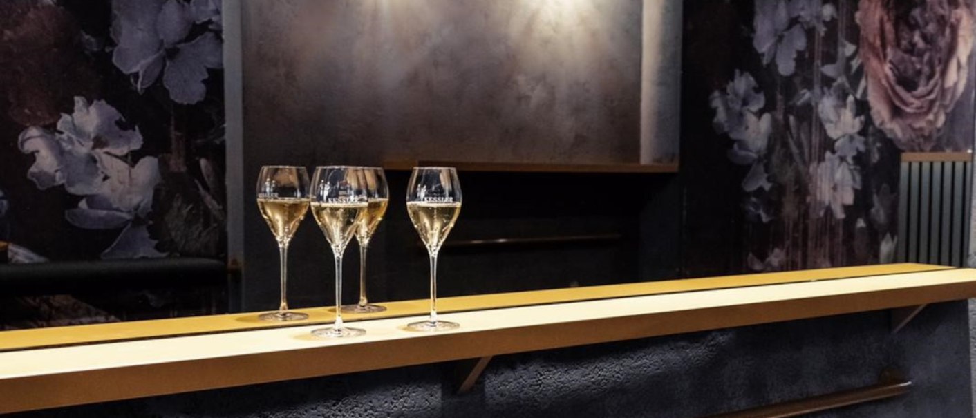 Drei Weingläser stehen auf einer Bar. Der Hintergrund ist dunkel mit floralen Mustern., © KESSLER Sekt GmbH & Co. KG