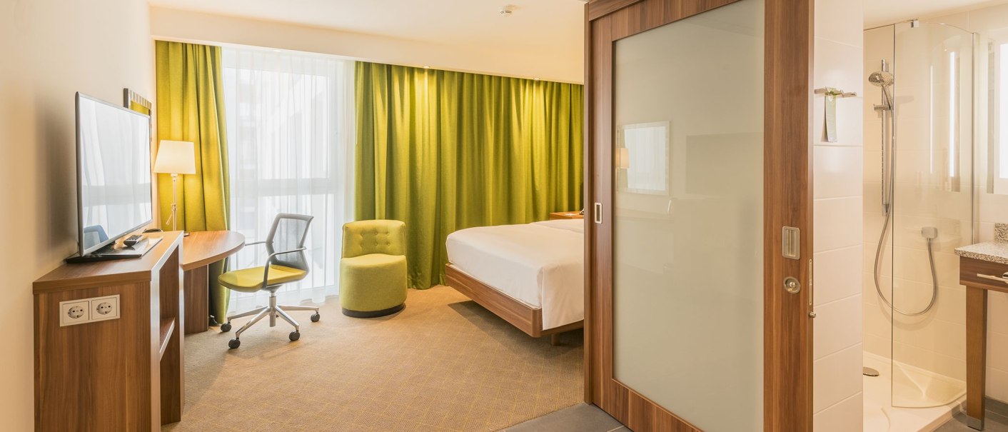 A modern hotel room with a double bed, a green armchair, a desk, and a view into the bathroom with a shower. Light-colored curtains and green drapes., &copy; Hampton by Hilton Stuttgart City Centre
