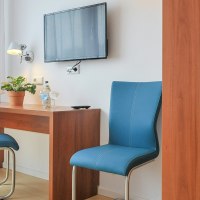 A simple hotel room with a desk, two blue chairs, a TV on the wall, and a closet., © Gästehaus ABZ Stuttgart A simple hotel room with a desk, two blue chairs, a TV on the wall, and a closet., © Gästehaus ABZ Stuttgart