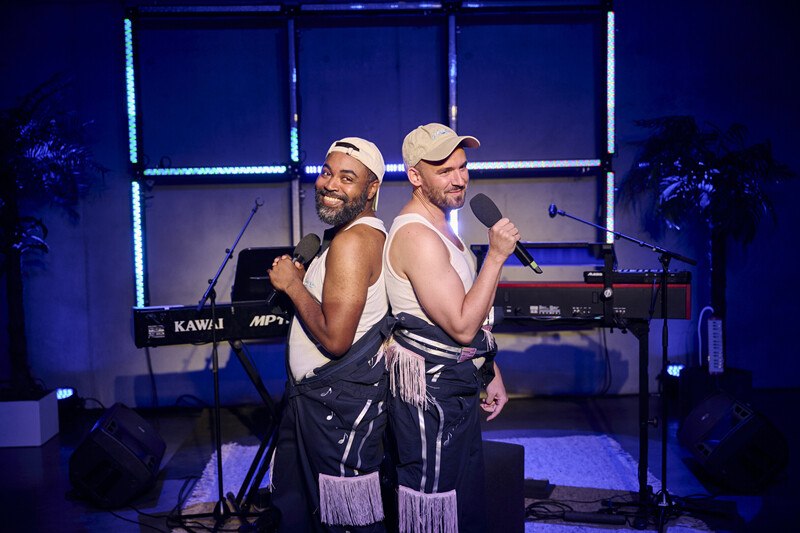 Zwei Männer stehen Rücken an Rücken auf einer Bühne, halten Mikrofone und lächeln. Im Hintergrund sind ein Keyboard und Palmen zu sehen., © Württembergische Staatstheater Stuttgart Zwei Männer stehen Rücken an Rücken auf einer Bühne, halten Mikrofone und lächeln. Im Hintergrund sind ein Keyboard und Palmen zu sehen., © Württembergische Staatstheater Stuttgart