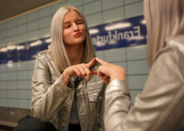 Frau mit silberner Jacke posiert vor einem Spiegel in einer U-Bahn-Station. Sie macht eine Handgeste und lächelt leicht., © Theaterhaus Stuttgart e.V. Frau mit silberner Jacke posiert vor einem Spiegel in einer U-Bahn-Station. Sie macht eine Handgeste und lächelt leicht., © Theaterhaus Stuttgart e.V.